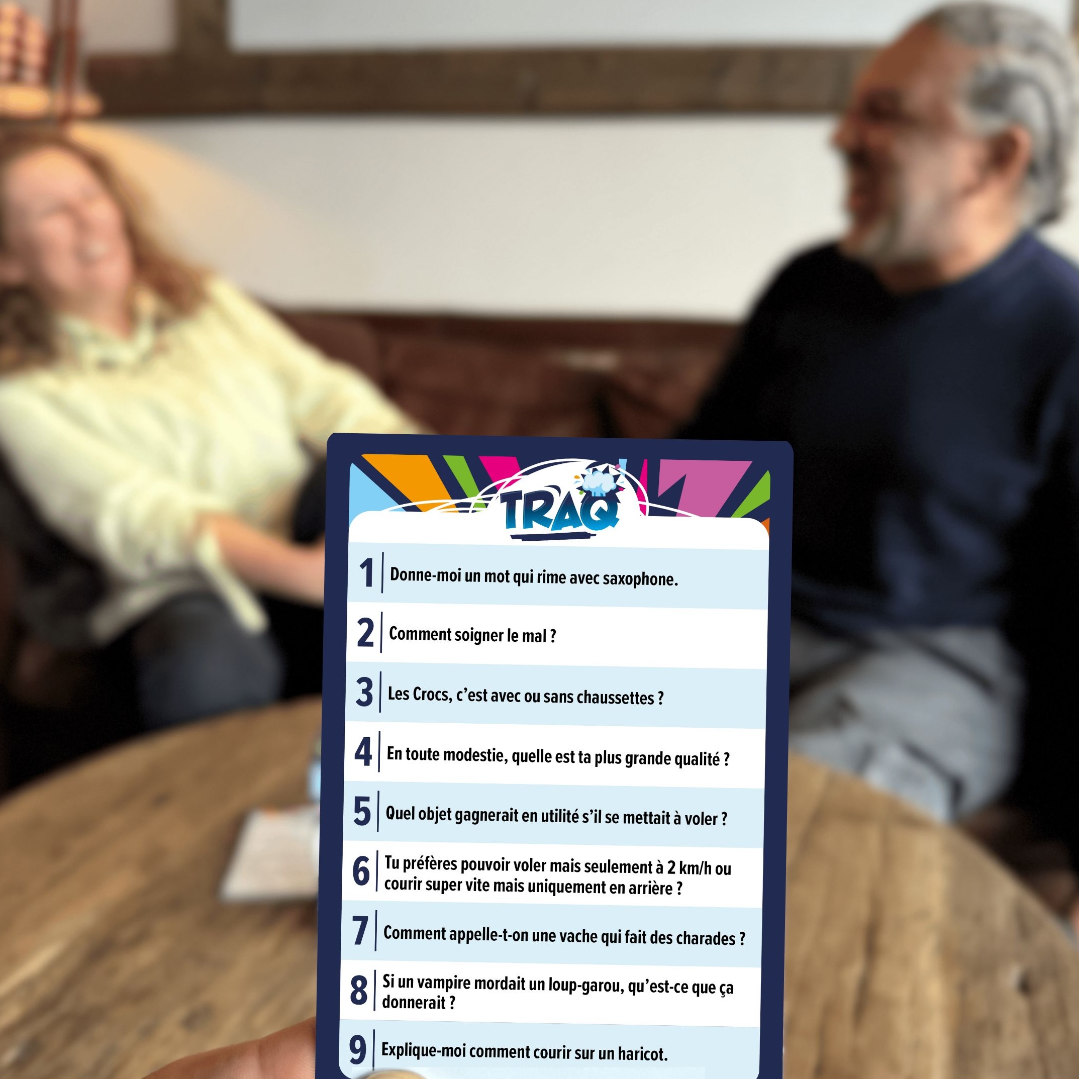 Deux adultes jouent au jeu de société TRAQ autour d'une table en bois, avec une carte de questions humoristiques au premier plan.