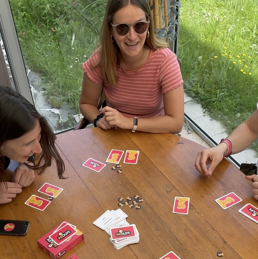Trois amies jouent au jeu de société Padupe autour d’une table en bois, en pleine partie, dans une véranda ensoleillée.