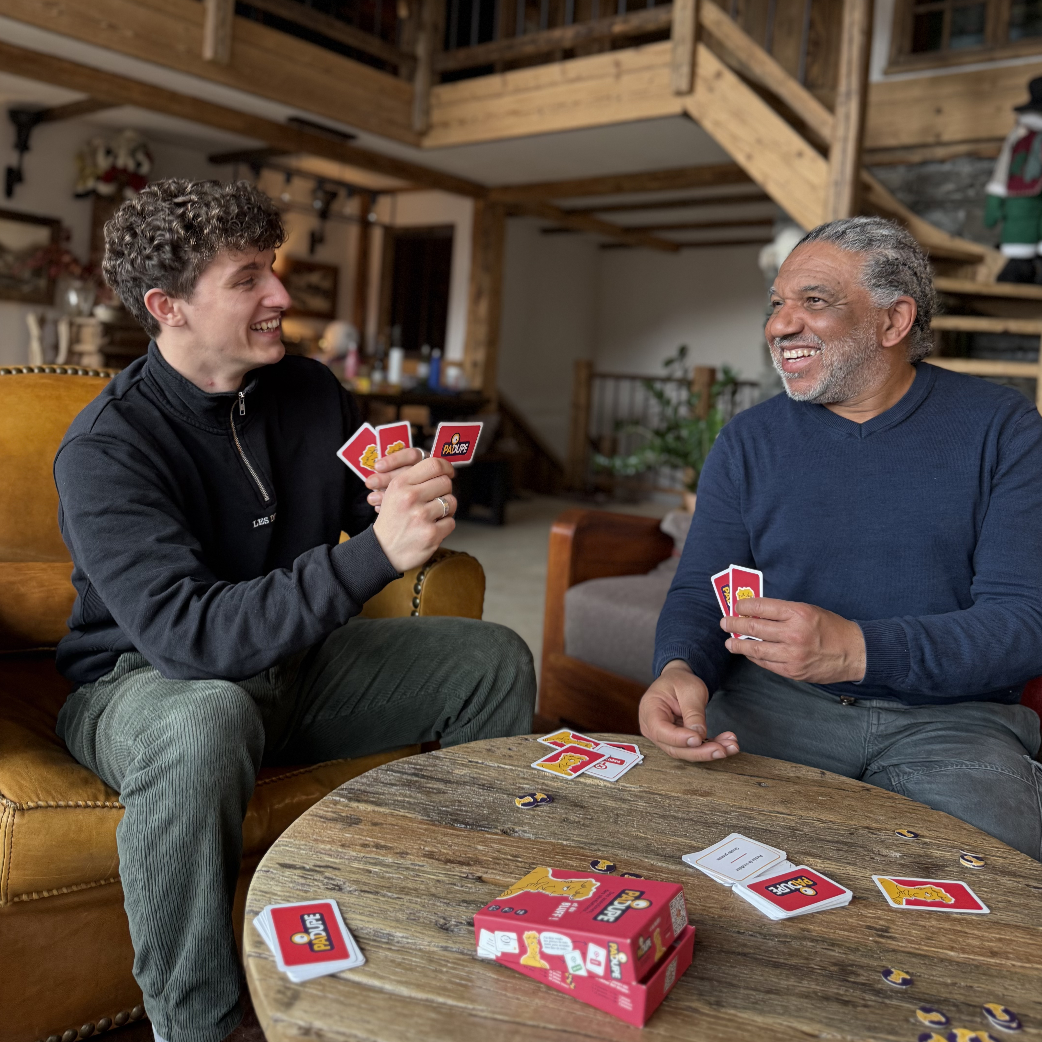 Deux hommes rient et jouent au jeu de société Padupe dans un chalet en bois, illustrant l'origine conviviale et artisanale du jeu.